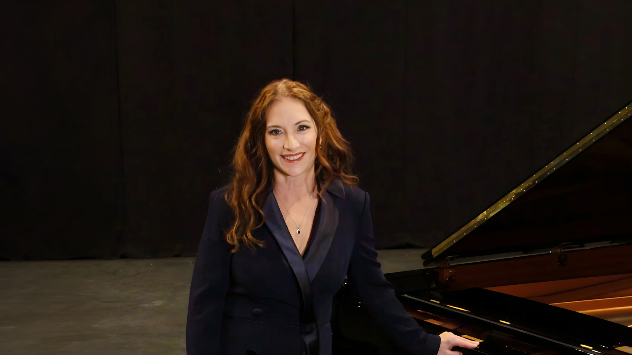 2627_Robin_WebHeader Robin Spielberg stands next to her piano smiling directly at the camera