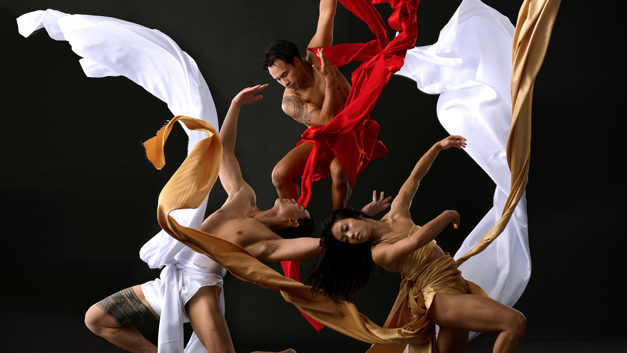 27_BlackGrace_WebHeader_1 Three performers from Black Grace pose with one another with one gracefully falling from above, all three wrapped in white, red, and gold cloth