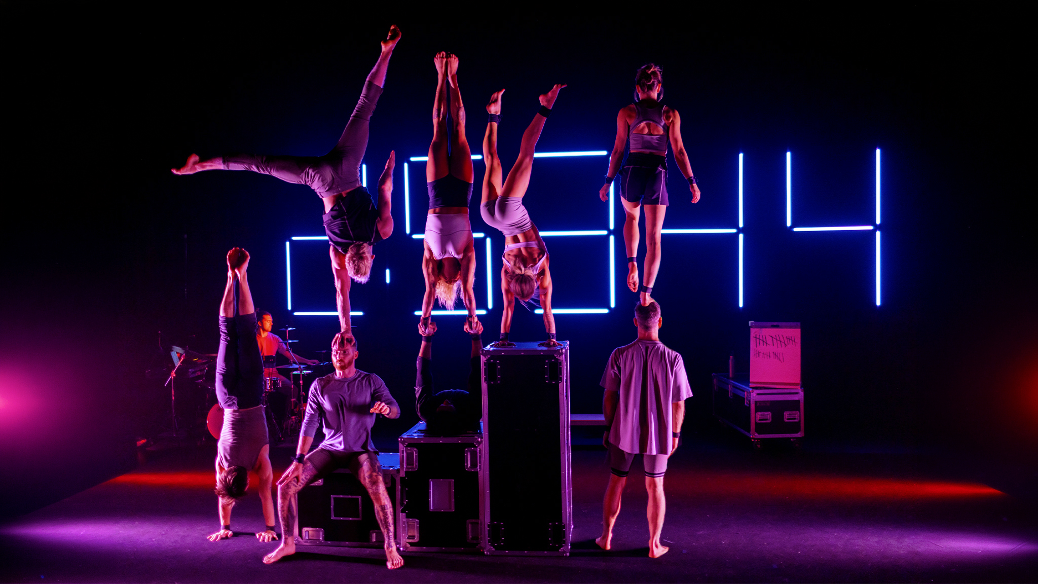 8 acrobats are seen on stage, half are balancing on other's heads while one is seen playing the drums, a large timer is seen behind them all